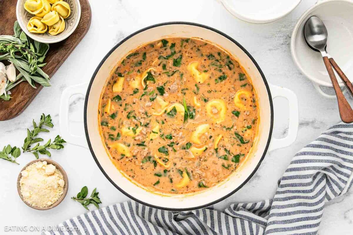 A white pot of Creamy Tortellini Soup with spinach sits on a white marble surface, surrounded by a bowl of cheese, fresh herbs, a striped towel, an empty bowl, and utensils.