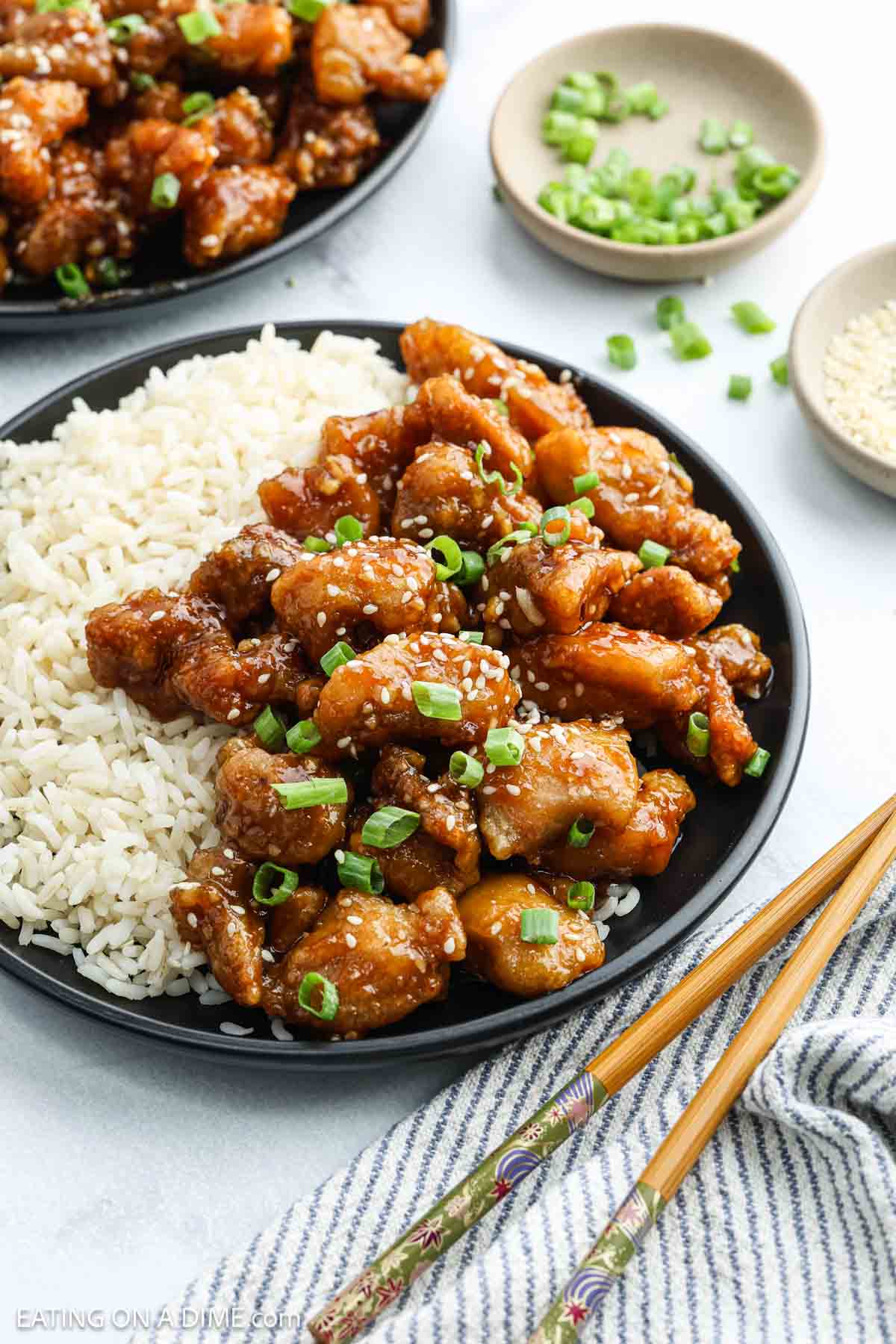 A black plate with white rice and crispy sesame chicken, inspired by classic Chinese chicken recipes, garnished with sesame seeds and green onions. Nearby are chopsticks, a blue-striped napkin, plus small bowls of green onions and sesame seeds.