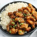 A black plate with white rice on one side and General Tso's Chicken—crispy pieces topped with sesame seeds and chopped green onions—on the other side.
