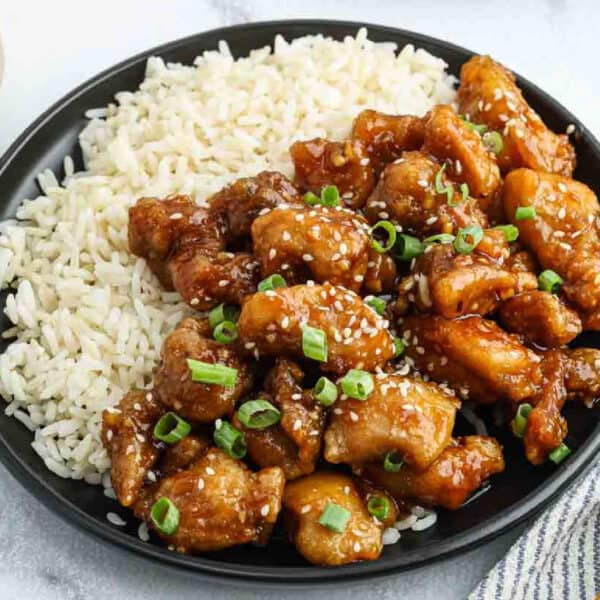A black plate with white rice on one side and General Tso's Chicken—crispy pieces topped with sesame seeds and chopped green onions—on the other side.