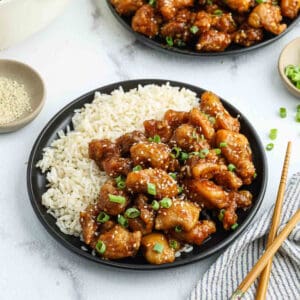 A black plate with white rice and pieces of glazed orange chicken, a classic of Chinese food, garnished with chopped green onions and sesame seeds, with chopsticks and small bowls of sesame seeds and green onions nearby.