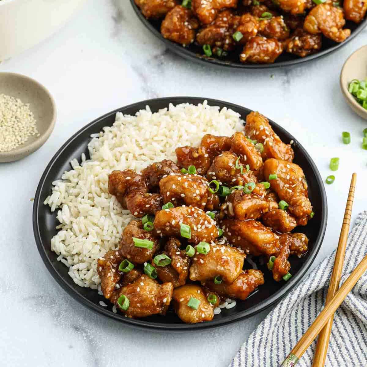A black plate with white rice and pieces of glazed orange chicken, a classic of Chinese food, garnished with chopped green onions and sesame seeds, with chopsticks and small bowls of sesame seeds and green onions nearby.