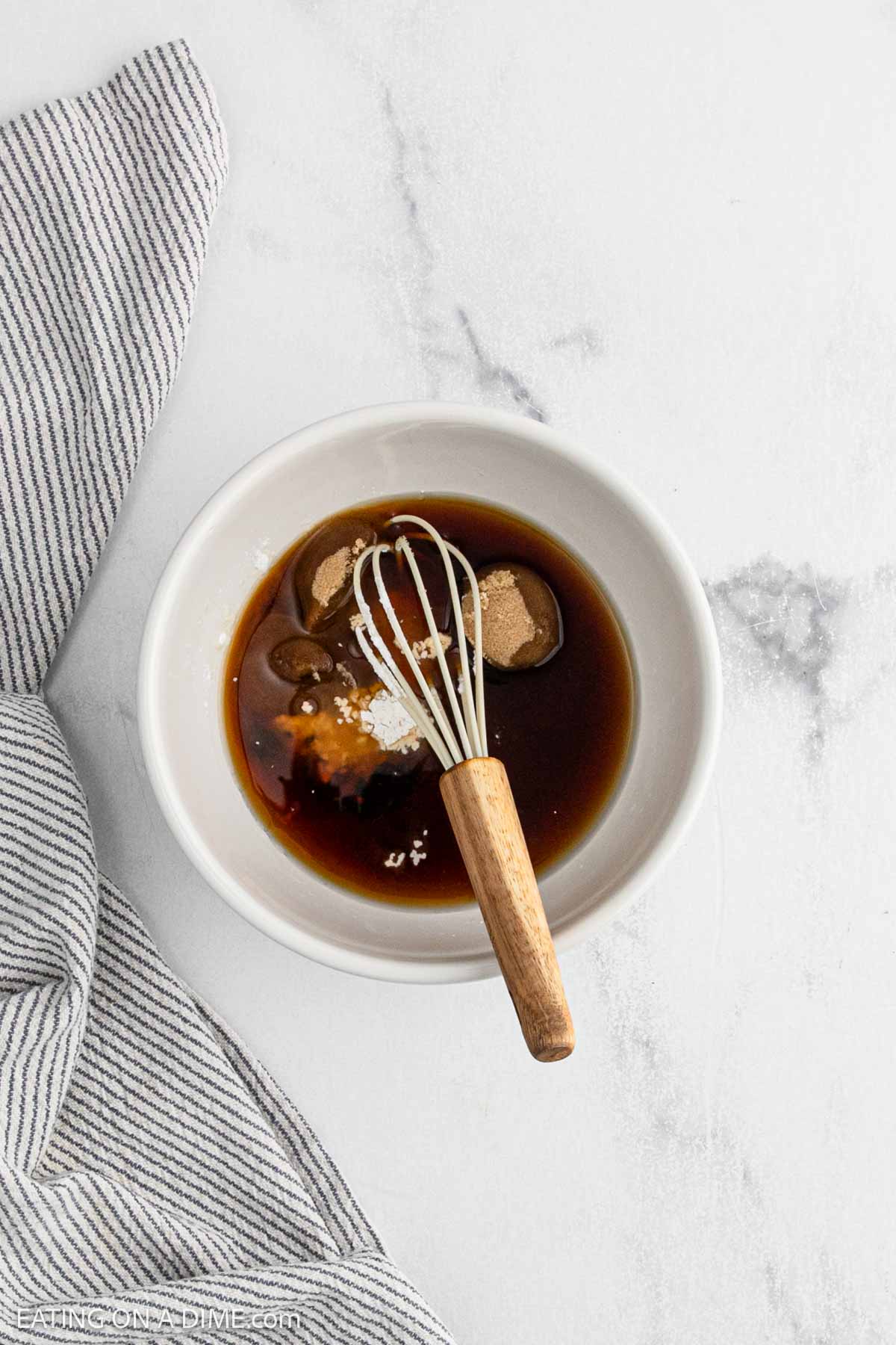 A white bowl with brown sauce, sugar, and spices being mixed by a small whisk with a wooden handle, perfect for an Asian cuisine dish like General Tso's Chicken, on a white marble surface next to a striped kitchen towel.