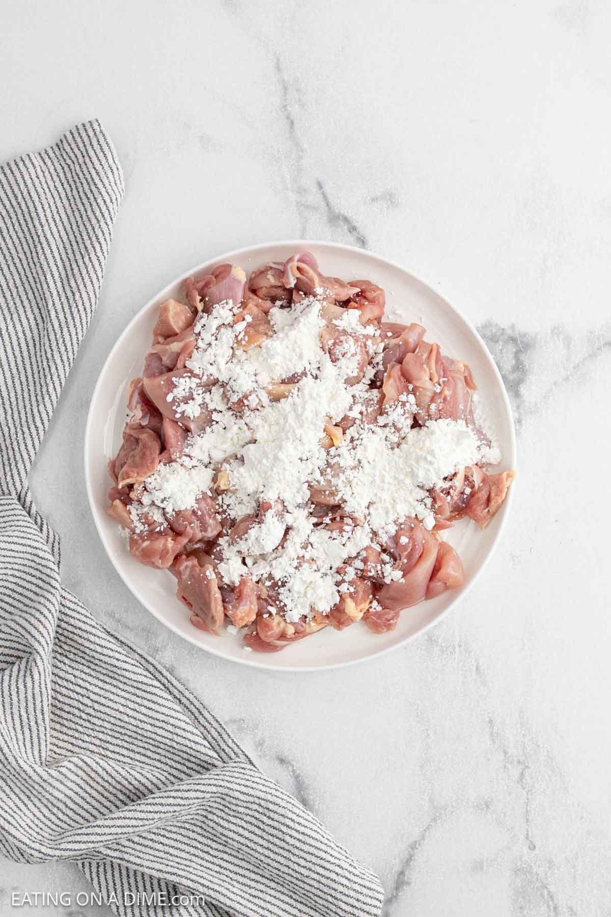 A white plate with raw, sliced chicken pieces topped with a pile of white flour sits on a light marble surface next to a gray and white striped cloth—a classic first step in preparing General Tso's Chicken, a favorite in Chinese food and Asian cuisine.