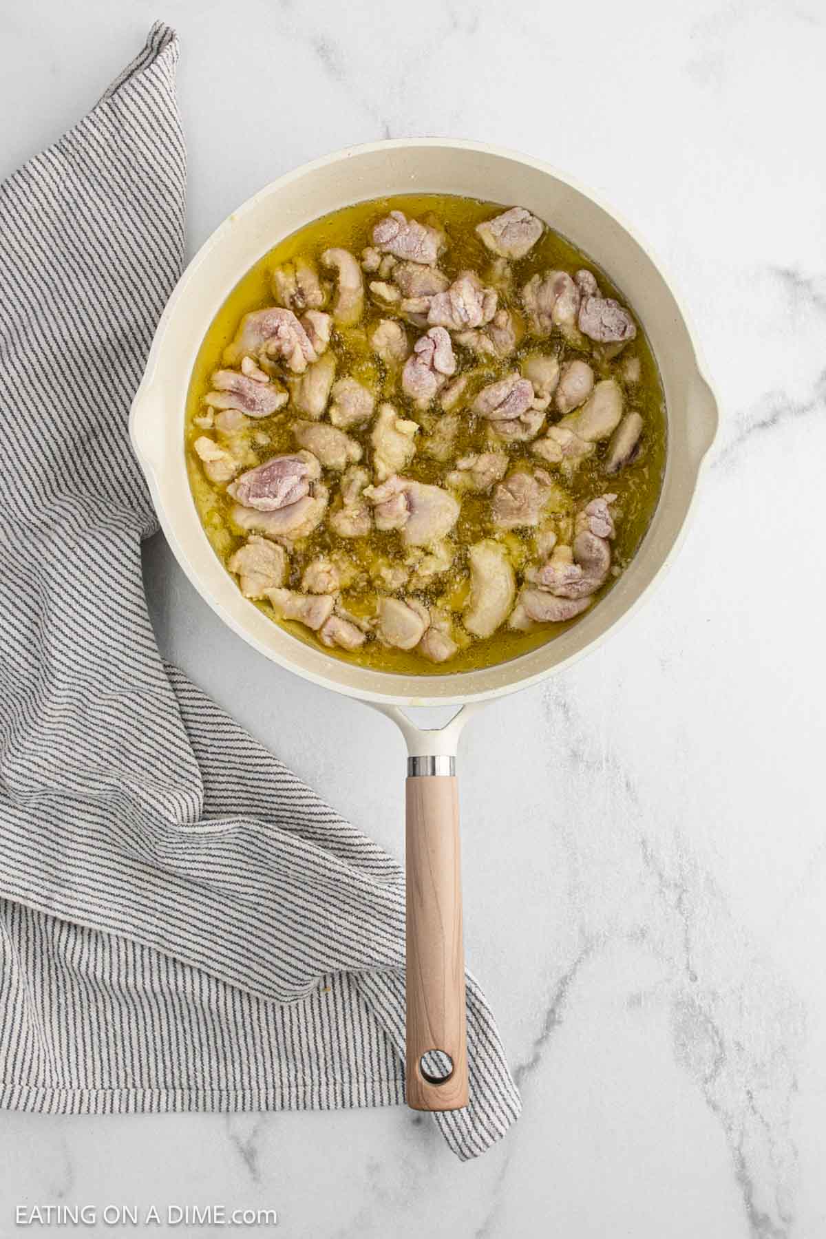 A white skillet filled with pieces of raw chicken cooking in olive oil sits on a white marble surface next to a folded gray and white striped cloth, ready for a delicious Chinese chicken recipe inspired by Asian cuisine.