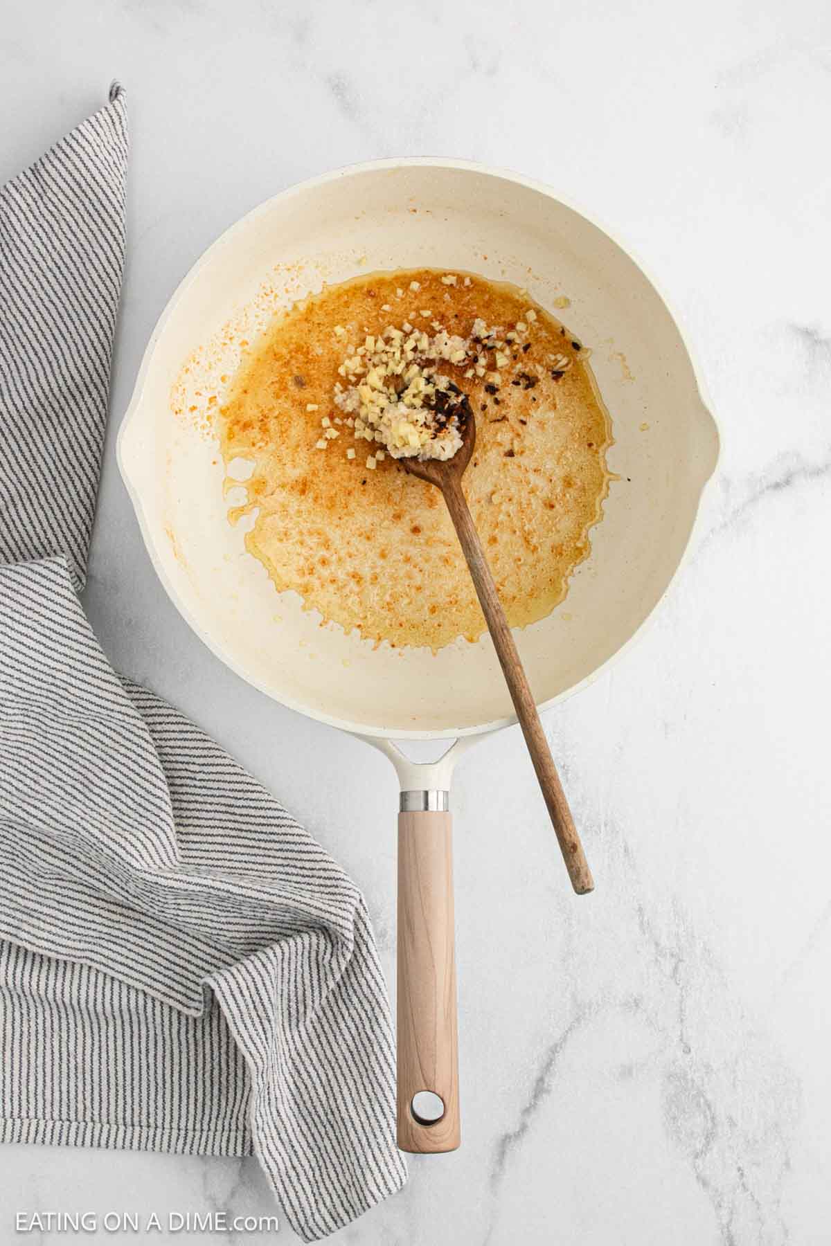 A white skillet with browned oil, minced garlic, and a wooden spoon rests on a marble counter next to a gray-striped kitchen towel—perfect prep for an easy chicken recipe inspired by classic Chinese food.