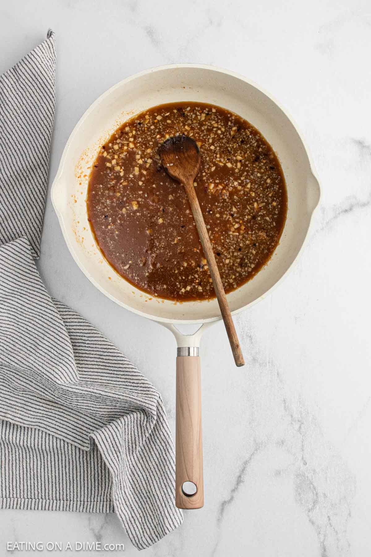 A white skillet filled with a brown, chunky General Tso's Chicken sauce and a wooden spoon resting inside, placed on a white marble surface next to a gray striped kitchen towel.