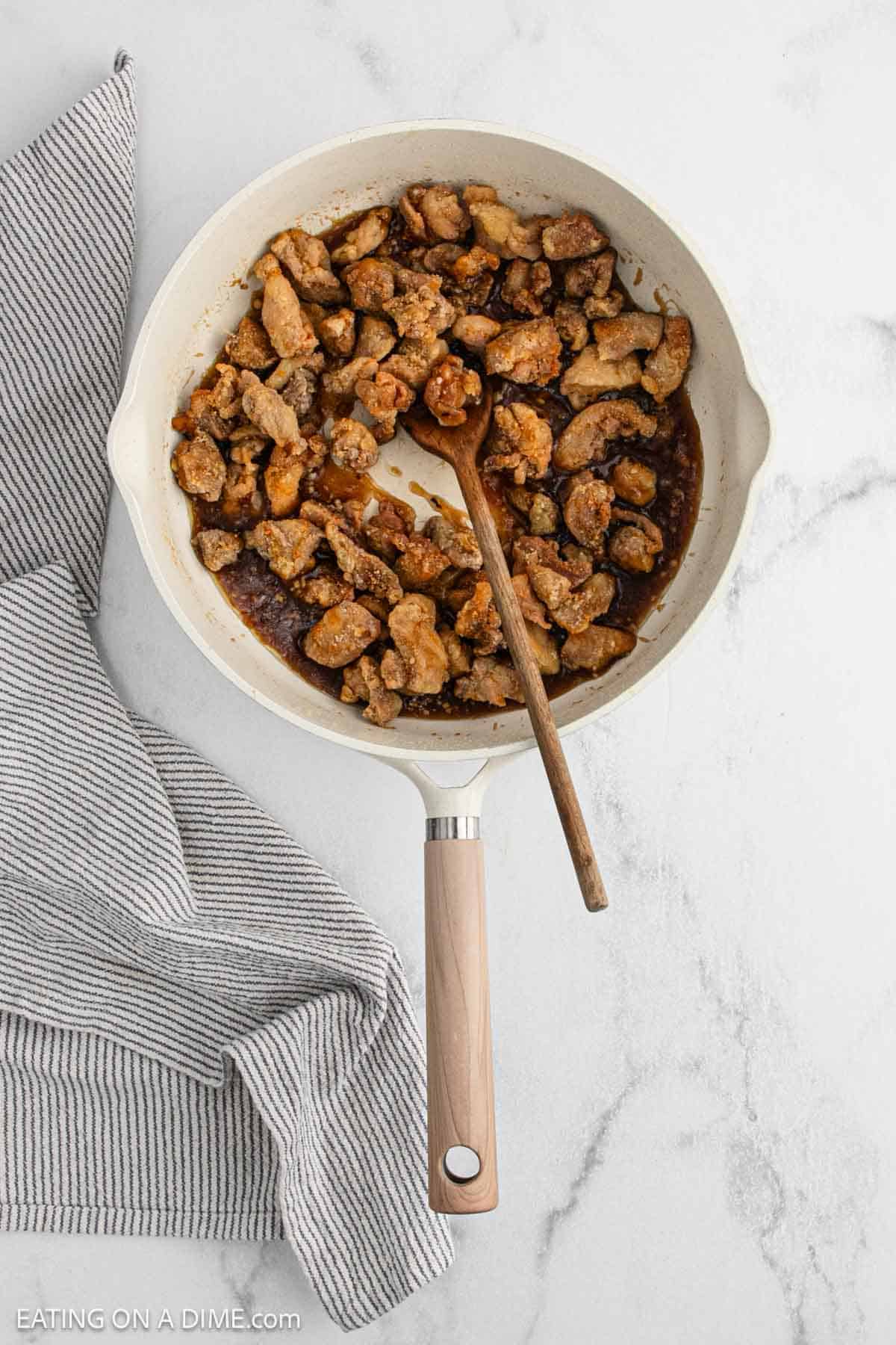 A white skillet filled with cooked, seasoned pieces of chicken, perfect for a homemade General Tso's Chicken recipe, with a wooden spoon resting inside. The skillet sits on a white marble surface next to a folded gray striped kitchen towel.