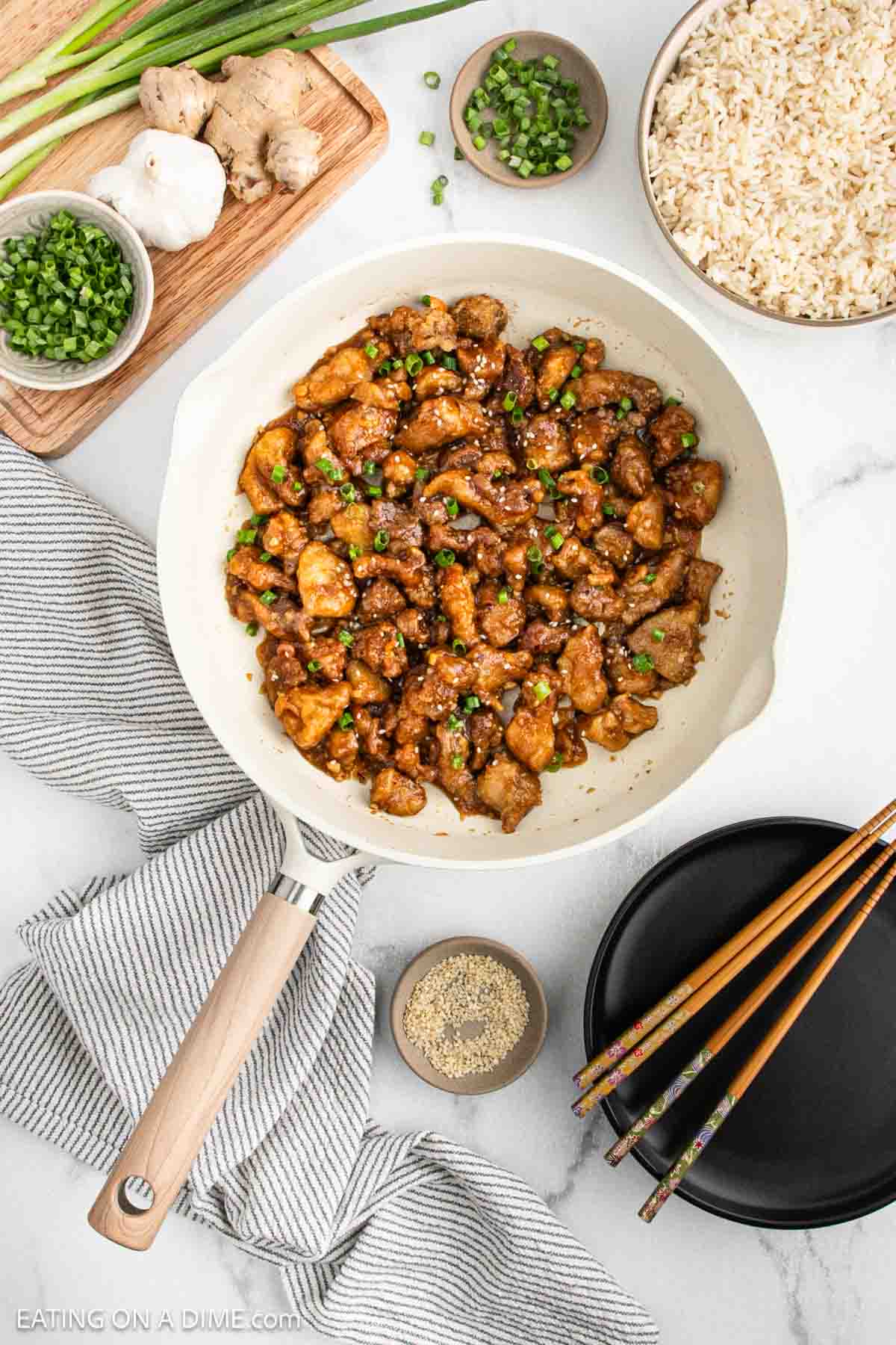A skillet filled with sesame chicken, inspired by a classic Chinese chicken recipe, topped with chopped green onions and surrounded by bowls of rice, sesame seeds, fresh ginger, and chopsticks on a white surface.