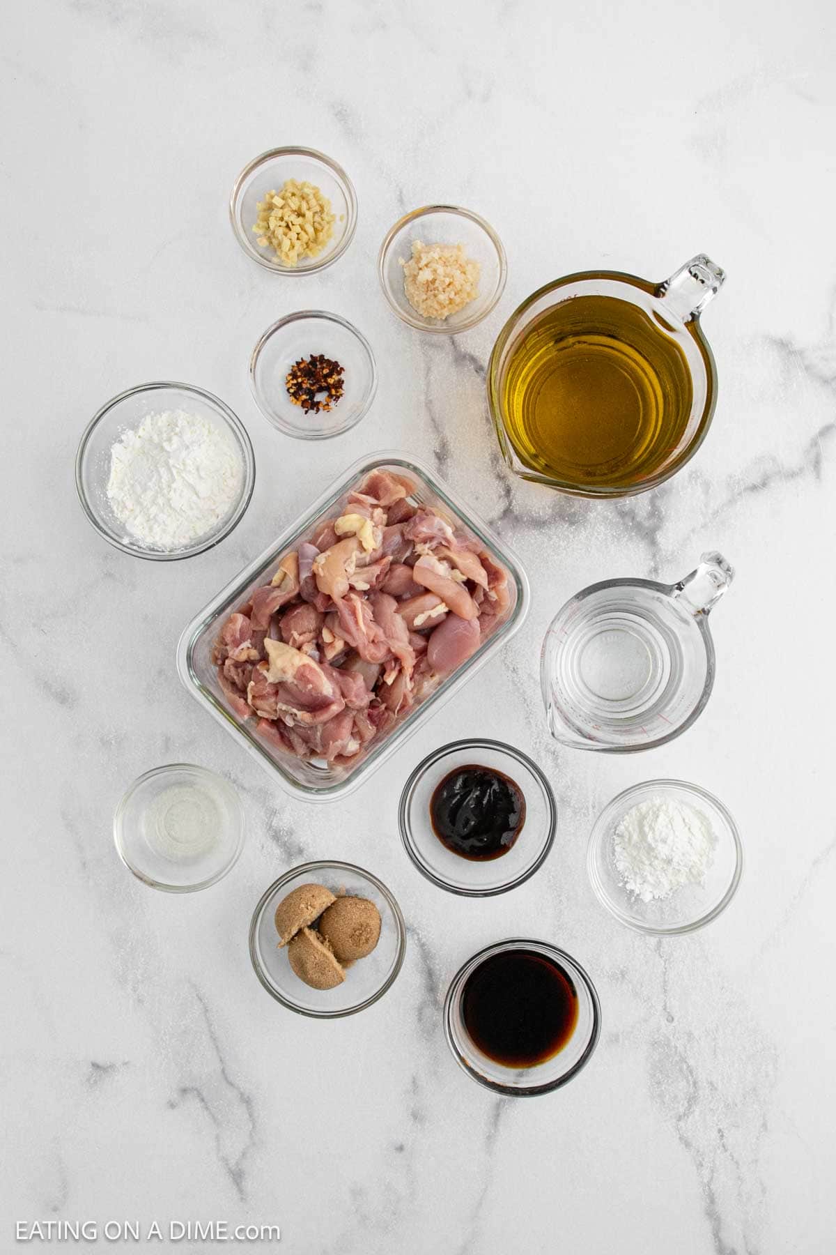 Overhead view of various ingredients in glass bowls on a marble surface, including raw chicken pieces, soy sauce, brown sugar, garlic, and seasonings—everything you need to make classic General Tso's Chicken from Asian cuisine.