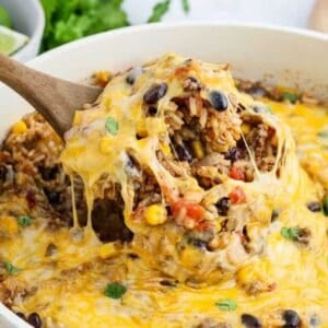 A wooden spoon scoops up a cheesy, baked Mexican casserole with melted cheese, black beans, corn, rice, and ground beef in a white dish. Perfect for Meal Plan 84, this dish is topped with fresh cilantro and diced tomatoes.