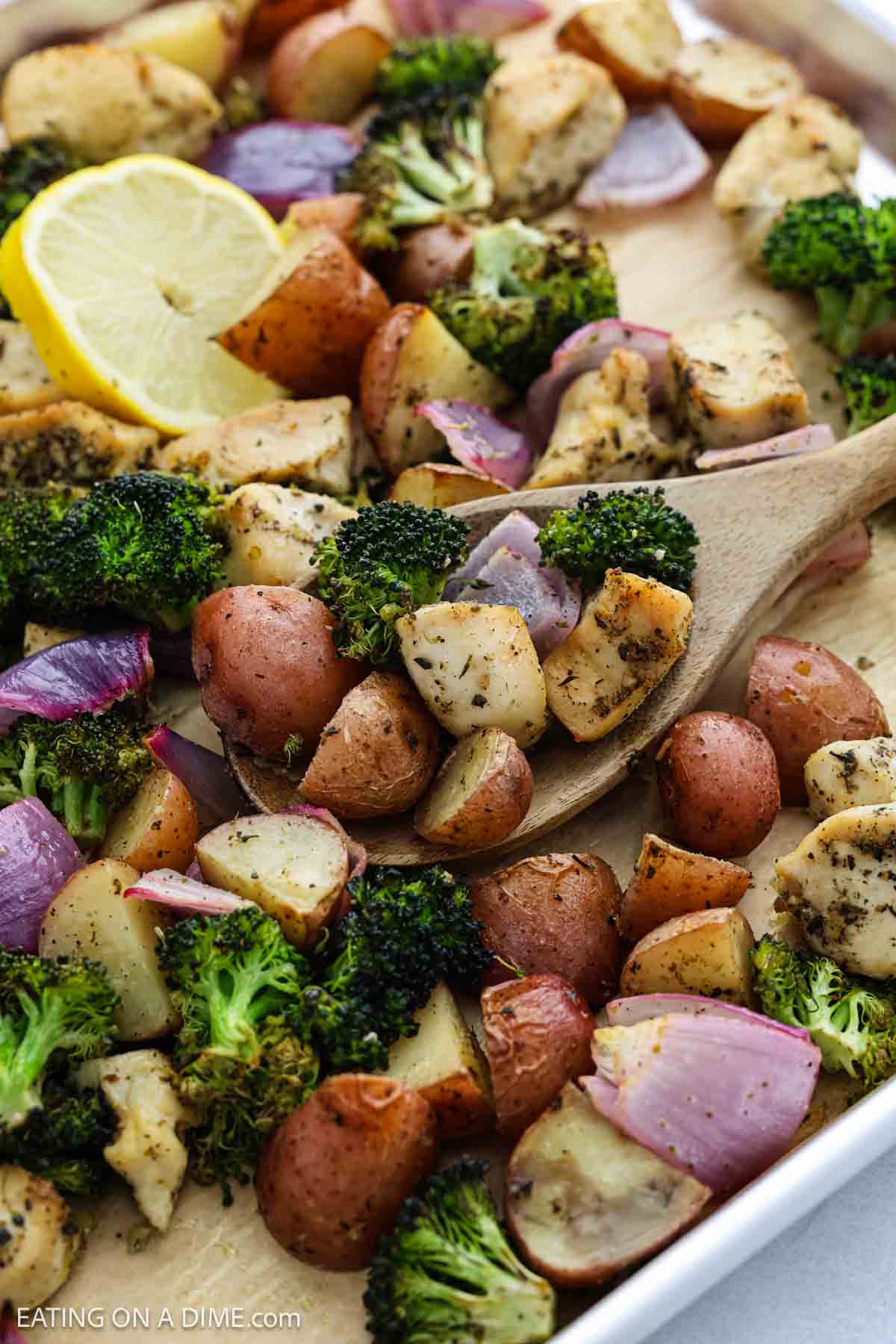 A sheet pan dinner featuring roasted red potatoes, broccoli, red onions, and seasoned chicken chunks, served with a wooden spoon and a lemon wedge on the side.