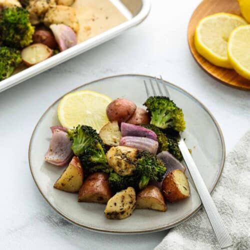 A plate with roasted potatoes, broccoli, and red onions, garnished with a lemon slice. A fork rests on the plate, with a tray of Sheet Pan Veggies and lemon slices on a wooden plate in the background.