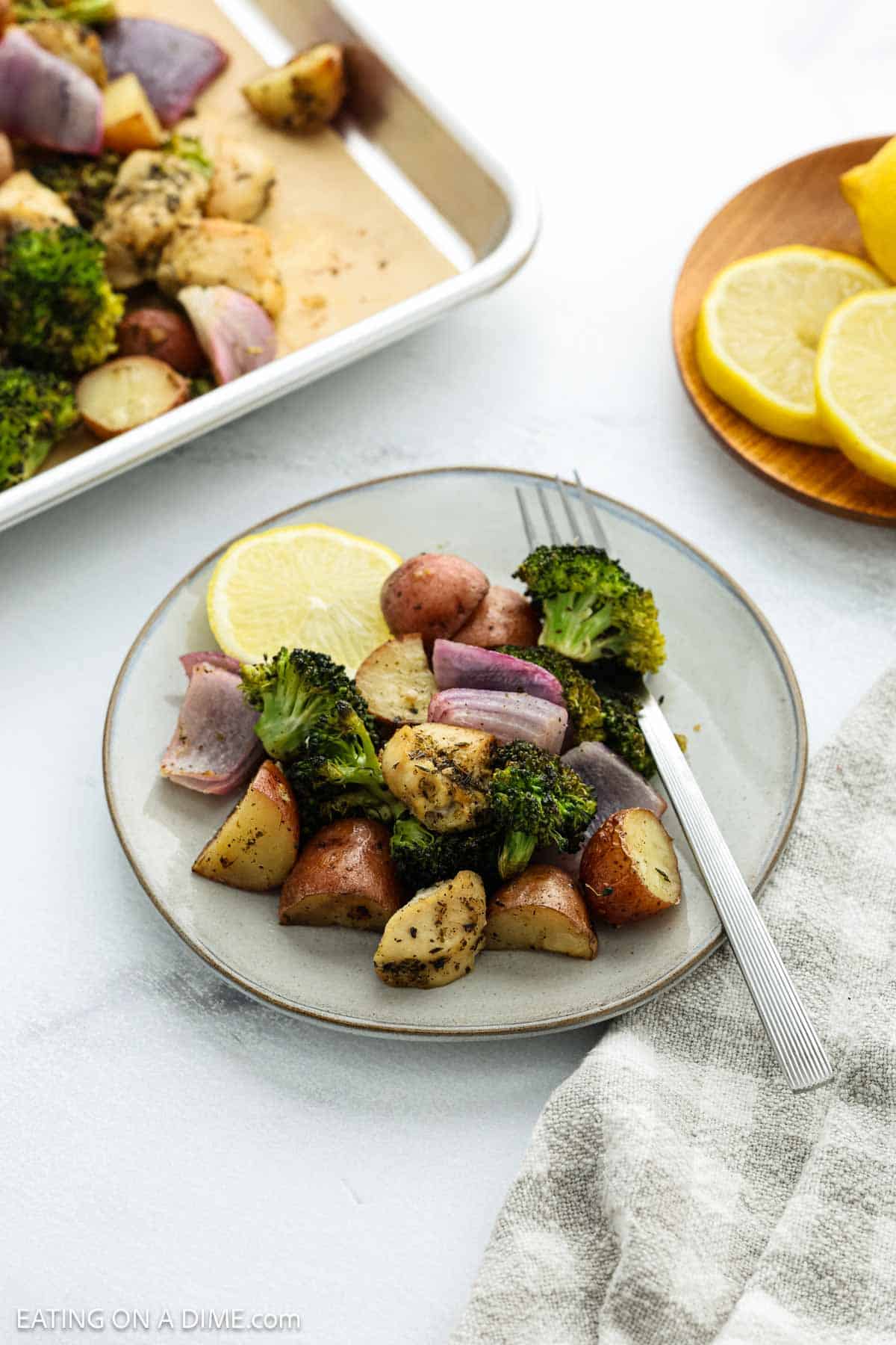 A vibrant sheet pan dinner of roasted vegetables—broccoli, red potatoes, and purple onions—garnished with a lemon slice. A fork rests on the plate, with extra veggies and a small plate of lemon slices in the background.