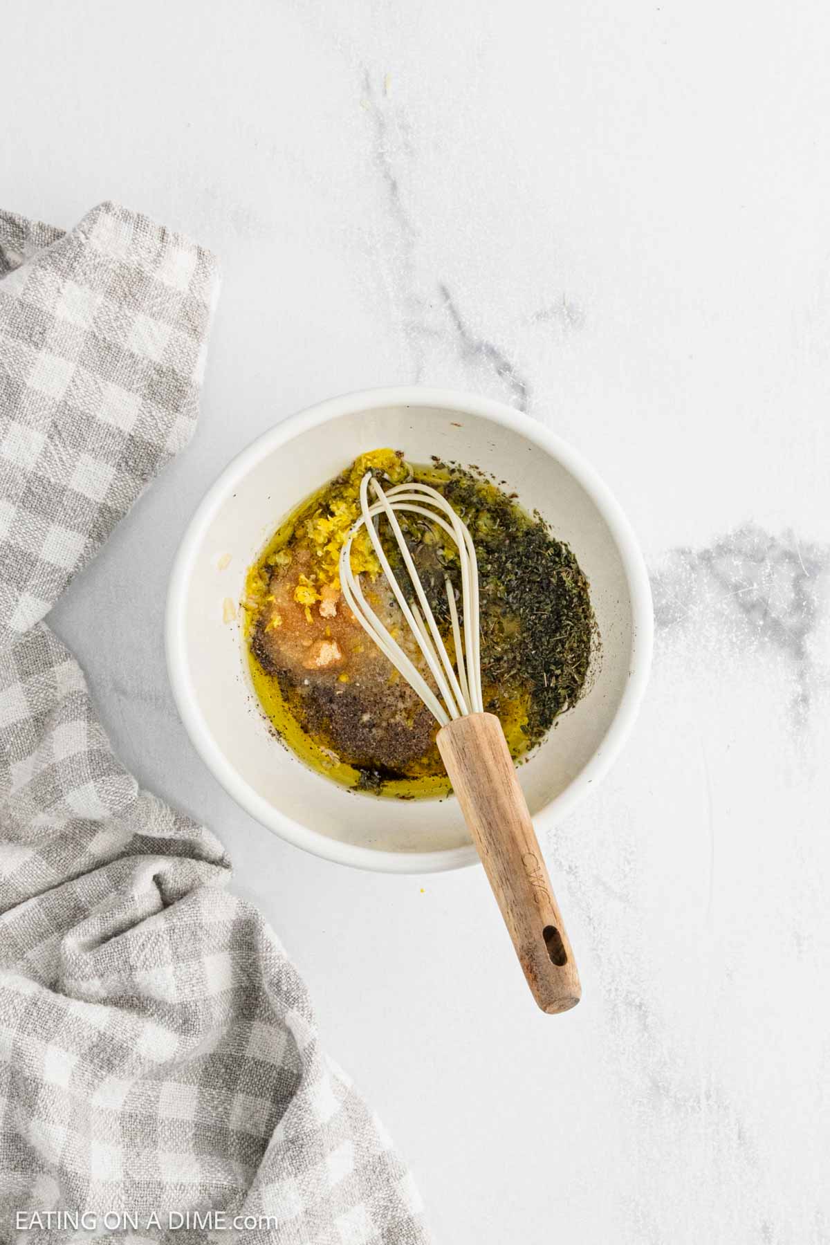 A white bowl with oil, spices, minced garlic, and a whisk inside sits on a white surface next to a gray and white checkered cloth—perfect for prepping your Sheet Pan Chicken and Veggies dinner.
