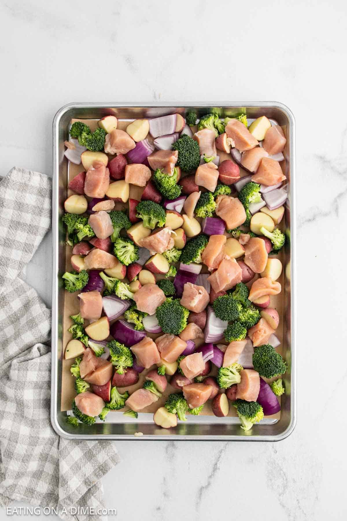 A baking sheet filled with chopped raw chicken and veggies—red potatoes, broccoli florets, red onion, and garlic cloves—ready for roasting. This easy Sheet Pan Chicken dinner sits beside a gray-and-white towel peeking in from the left side.