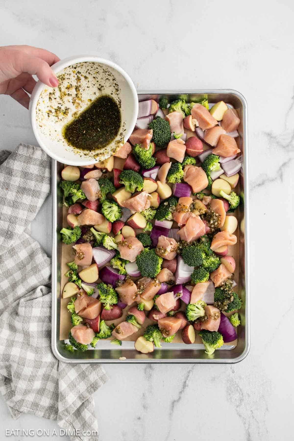 A hand pours a bowl of herb marinade over a sheet pan filled with chopped chicken and veggies—broccoli, red onion, potatoes, and garlic cloves. A gray checked towel sits nearby on the marble countertop.