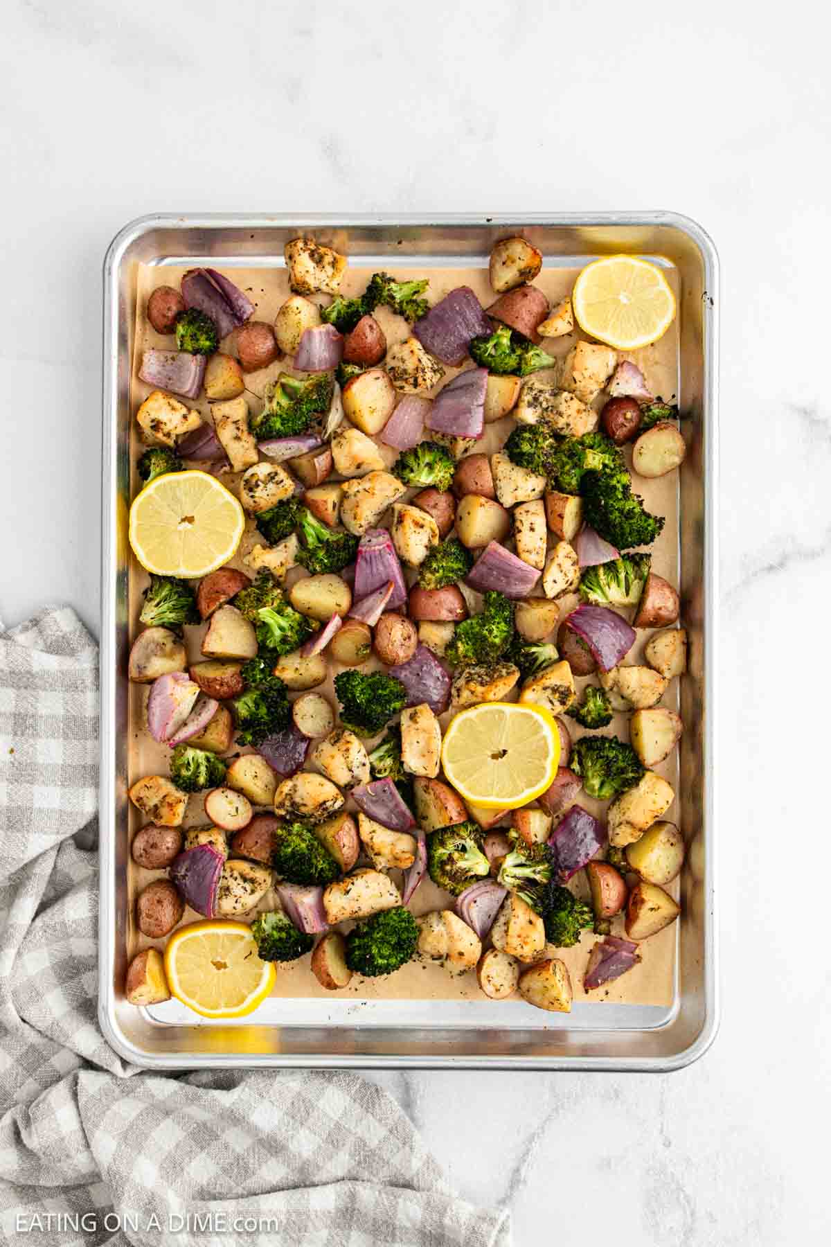 A sheet pan filled with roasted diced potatoes, broccoli florets, red onion chunks, and chicken pieces makes this Sheet Pan Chicken and Veggies meal pop, garnished with lemon slices. The pan rests on a light surface beside a striped cloth.