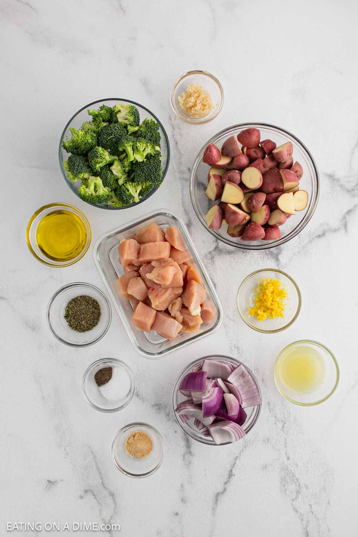 An overhead view of separated ingredients for Sheet Pan Chicken and Veggies on a white surface: chopped broccoli, halved red potatoes, diced raw chicken, red onion chunks, minced garlic, olive oil, lemon zest and juice, plus various seasonings in small bowls.