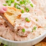 A hand dips a piece of bread into a creamy Shrimp Dip seafood salad topped with chopped green onions, in a white bowl.