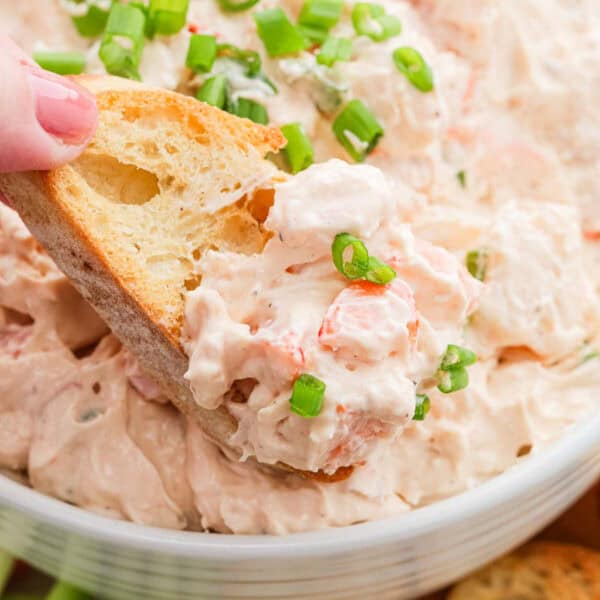 A hand dips a piece of bread into a creamy Shrimp Dip seafood salad topped with chopped green onions, in a white bowl.