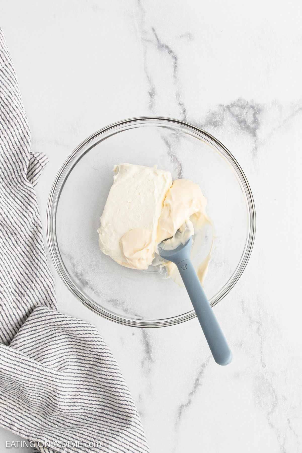A glass bowl with cream cheese and a blue spatula sits on a white marble surface next to a gray and white striped cloth.