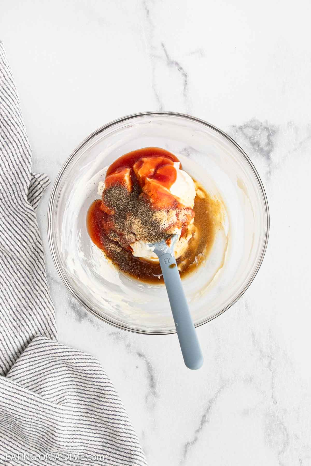 A glass bowl with mayonnaise, ketchup, black pepper, and Worcestershire sauce being mixed with a blue spatula, placed on a marble surface next to a striped cloth.