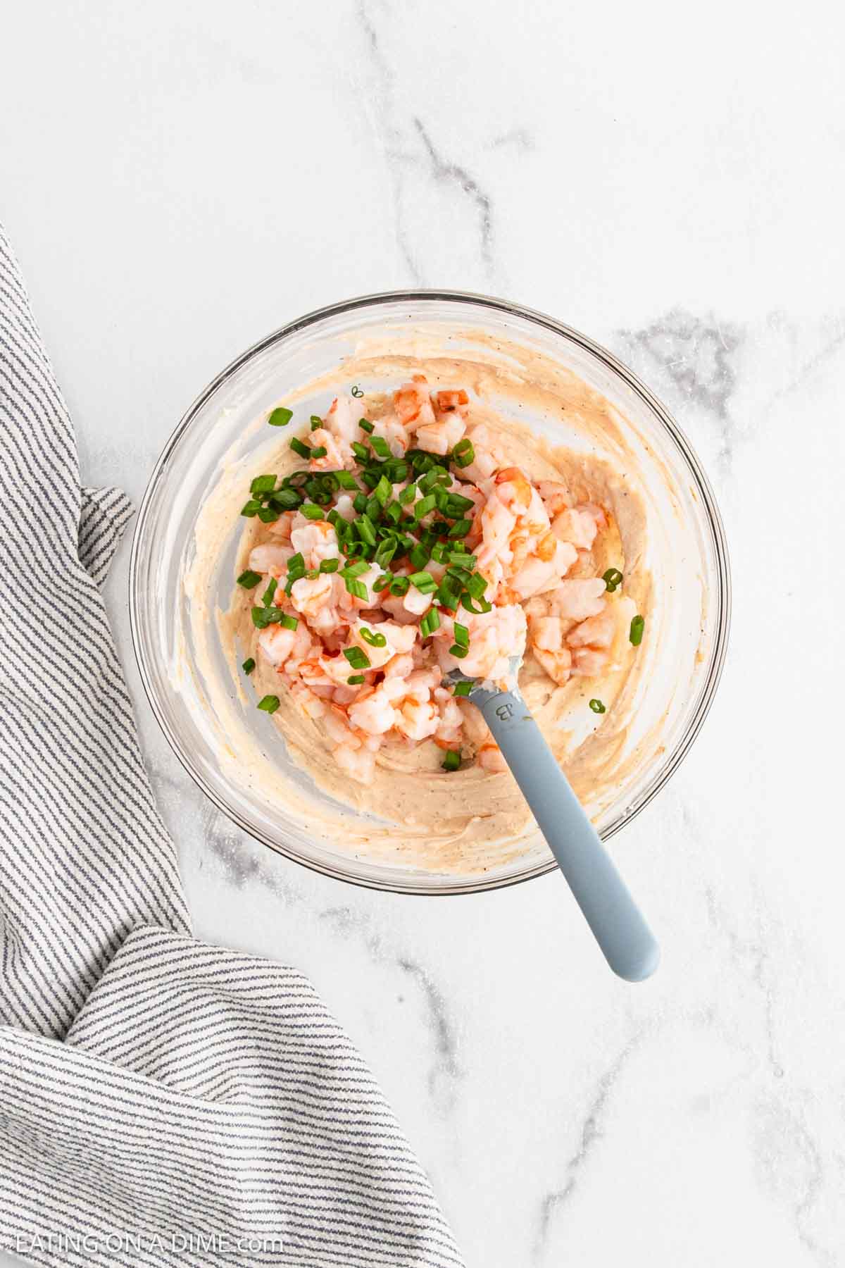 A glass bowl filled with a creamy mixture, diced shrimp, and chopped chives, with a light blue spatula resting inside. The bowl sits on a white marble surface next to a striped cloth.