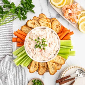 A bowl of creamy shrimp dip topped with chopped green onions, surrounded by toasted bread slices, celery, and carrot sticks on a marble surface. Lemon slices, shrimp, and fresh parsley are arranged nearby.