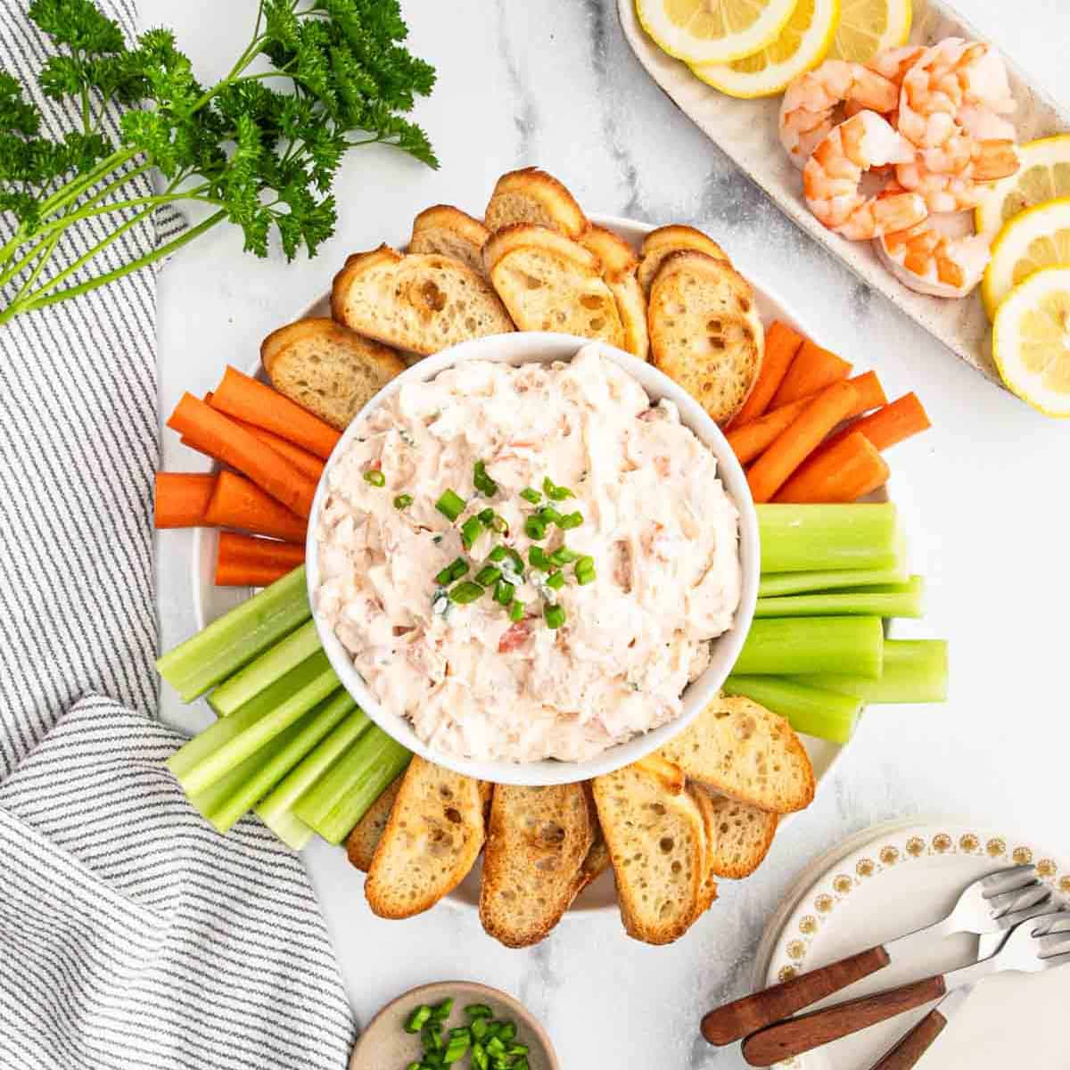 A bowl of creamy shrimp dip topped with chopped green onions, surrounded by toasted bread slices, celery, and carrot sticks on a marble surface. Lemon slices, shrimp, and fresh parsley are arranged nearby.