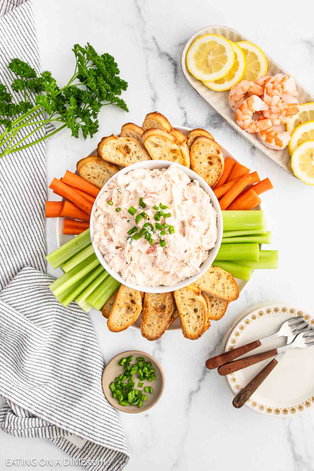 A bowl of shrimp dip is surrounded by crostini, celery, and carrot sticks on a platter. Nearby are lemon slices, shrimp, chopped green onions, parsley, napkins, and utensils on a marble surface.