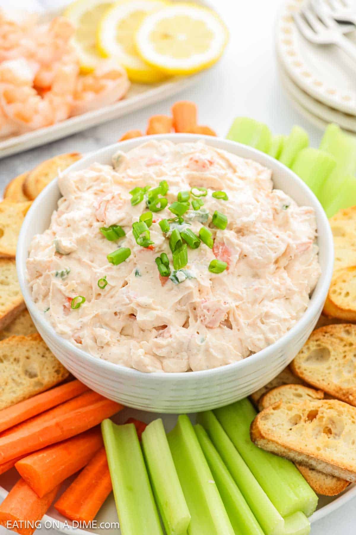 A bowl of creamy shrimp dip topped with chopped green onions, surrounded by sliced carrots, celery sticks, and toasted baguette slices on a white platter. Lemon slices and shrimp are visible in the background.