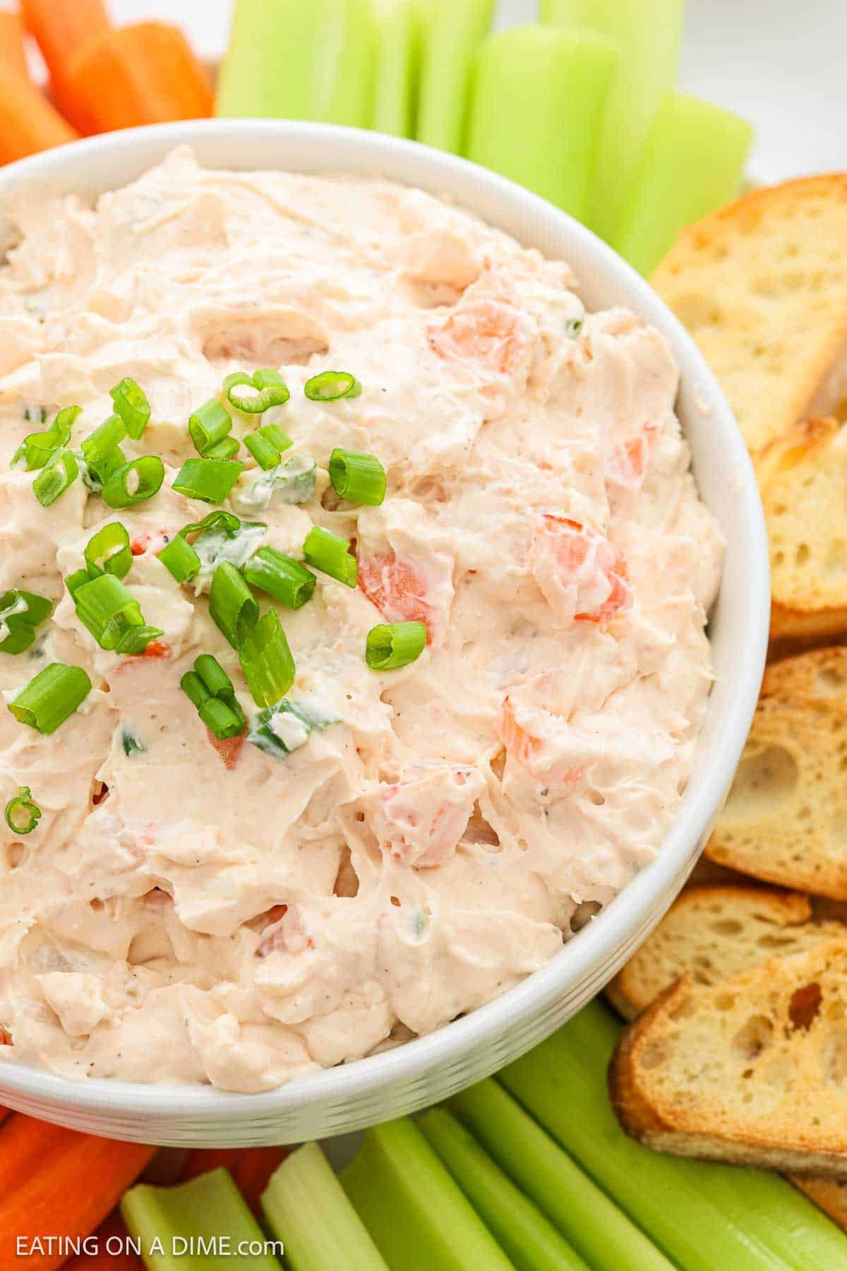 A bowl of creamy dip garnished with chopped green onions, surrounded by celery sticks, carrot sticks, and toasted bread slices.