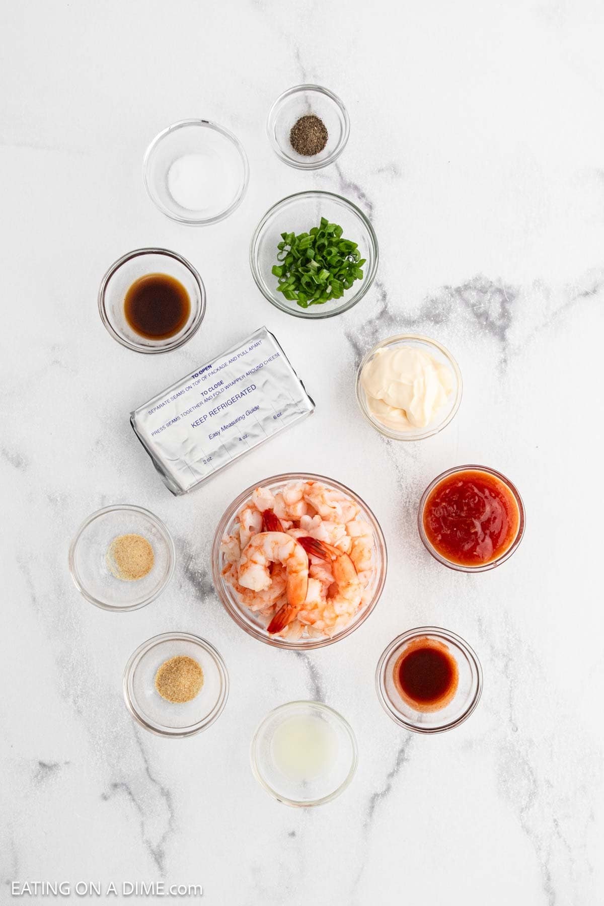 Overhead view of ingredients in small bowls on a white surface, including cooked shrimp, cream cheese, mayonnaise, cocktail sauce, green onions, lemon juice, seasonings, and various sauces.
