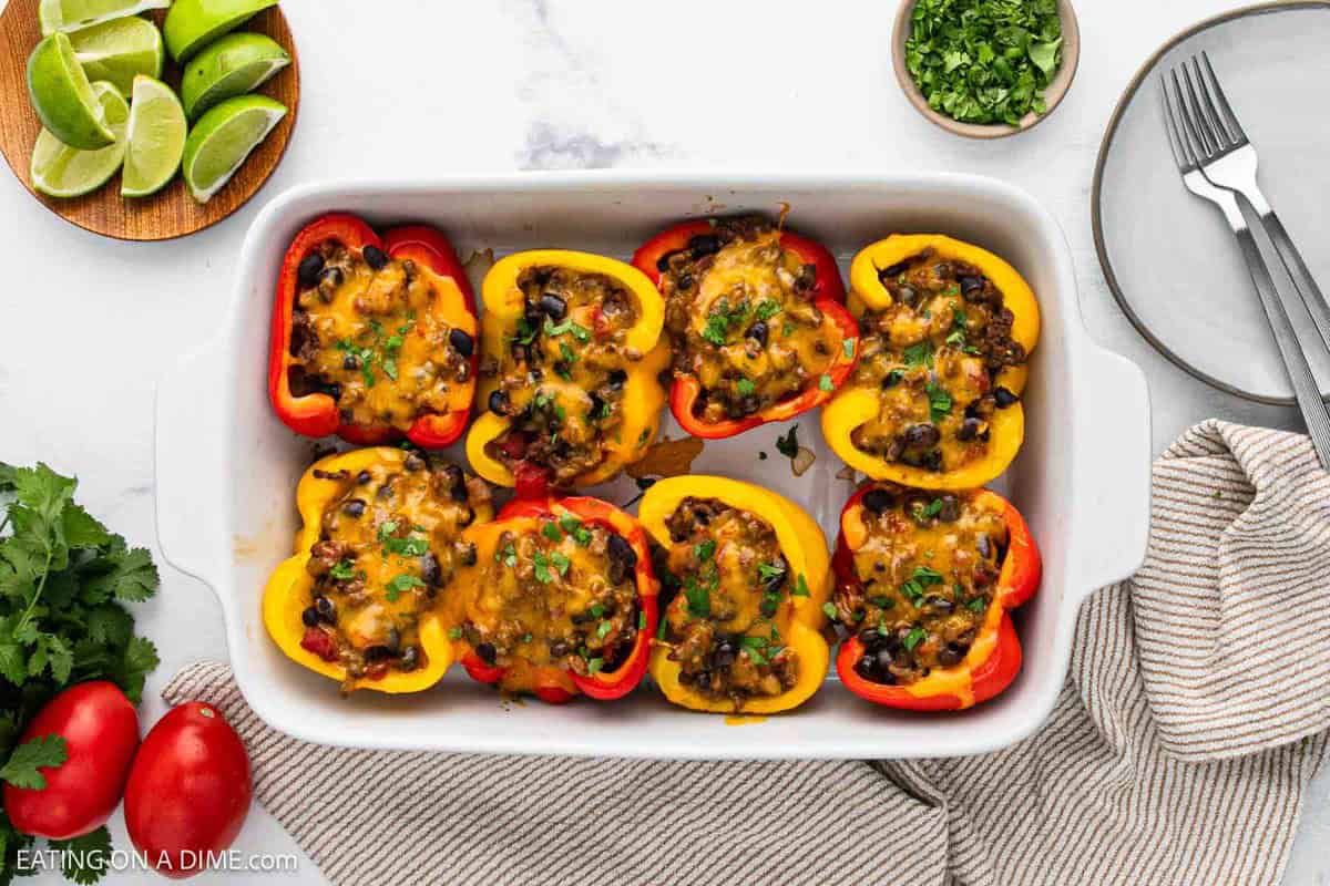 A white baking dish filled with Taco Stuffed Peppers—halved red and yellow bell peppers packed with a black bean, corn, and cheese mixture, garnished with herbs. Plates, limes, tomatoes, and cilantro surround this vibrant taco recipe.