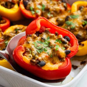 Close-up of Taco Stuffed Peppers filled with a cheesy mixture of ground meat, beans, and herbs, garnished with chopped cilantro in a white baking dish. A red pepper is being lifted out with a serving spoon—perfect for any taco recipe night.