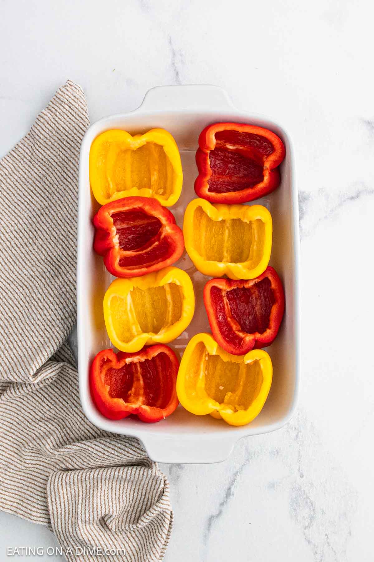 A white baking dish holds eight taco stuffed peppers—four red and four yellow—cut side up, beside a beige striped kitchen towel on a white marble surface.