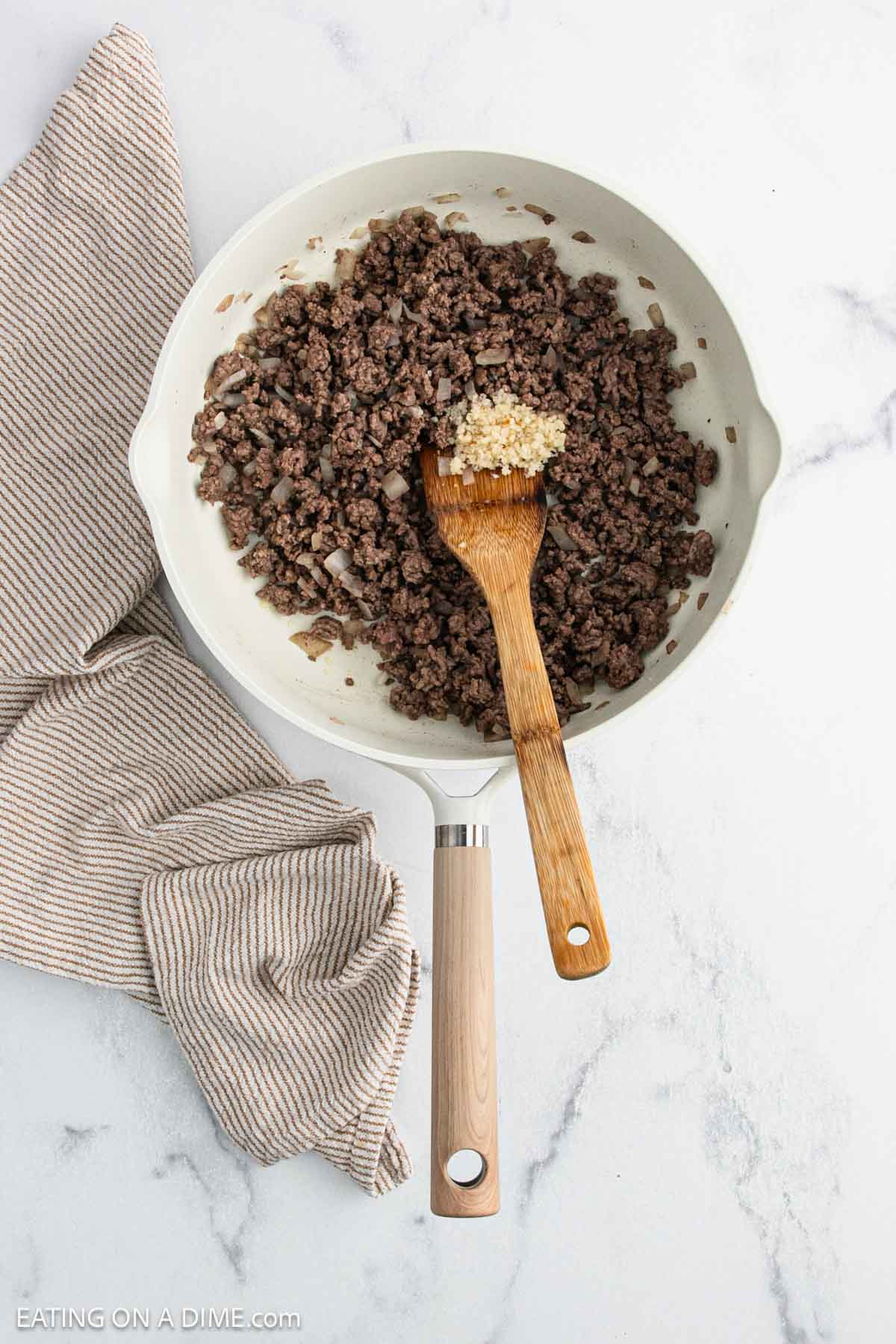 A white skillet with cooked ground beef and minced garlic for Taco Stuffed Peppers being stirred with a wooden spoon, next to a beige-striped kitchen towel on a marble countertop.
