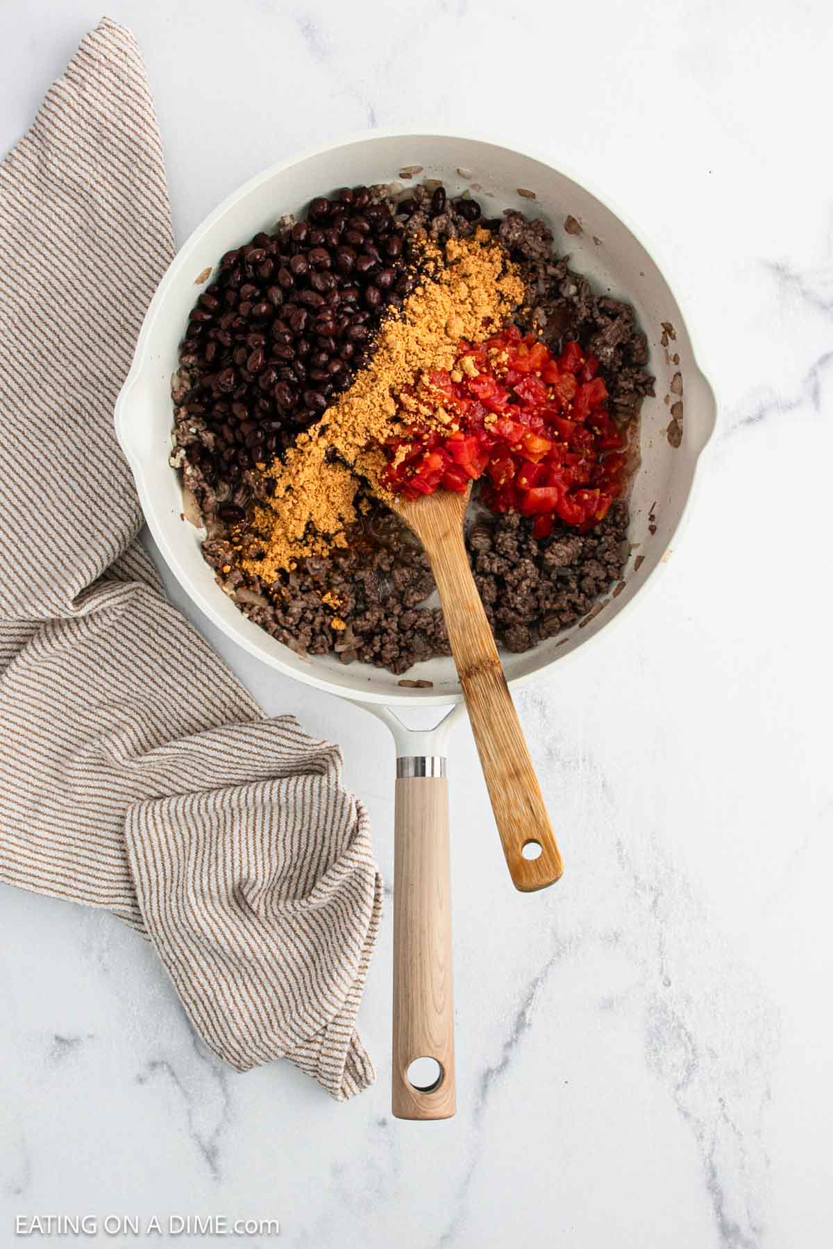 A skillet on a white marble surface holds cooked ground beef, black beans, taco seasoning, and diced tomatoes—perfect for a Taco Stuffed Peppers taco recipe. A wooden spoon rests inside, with a beige-striped kitchen towel beside the pan.