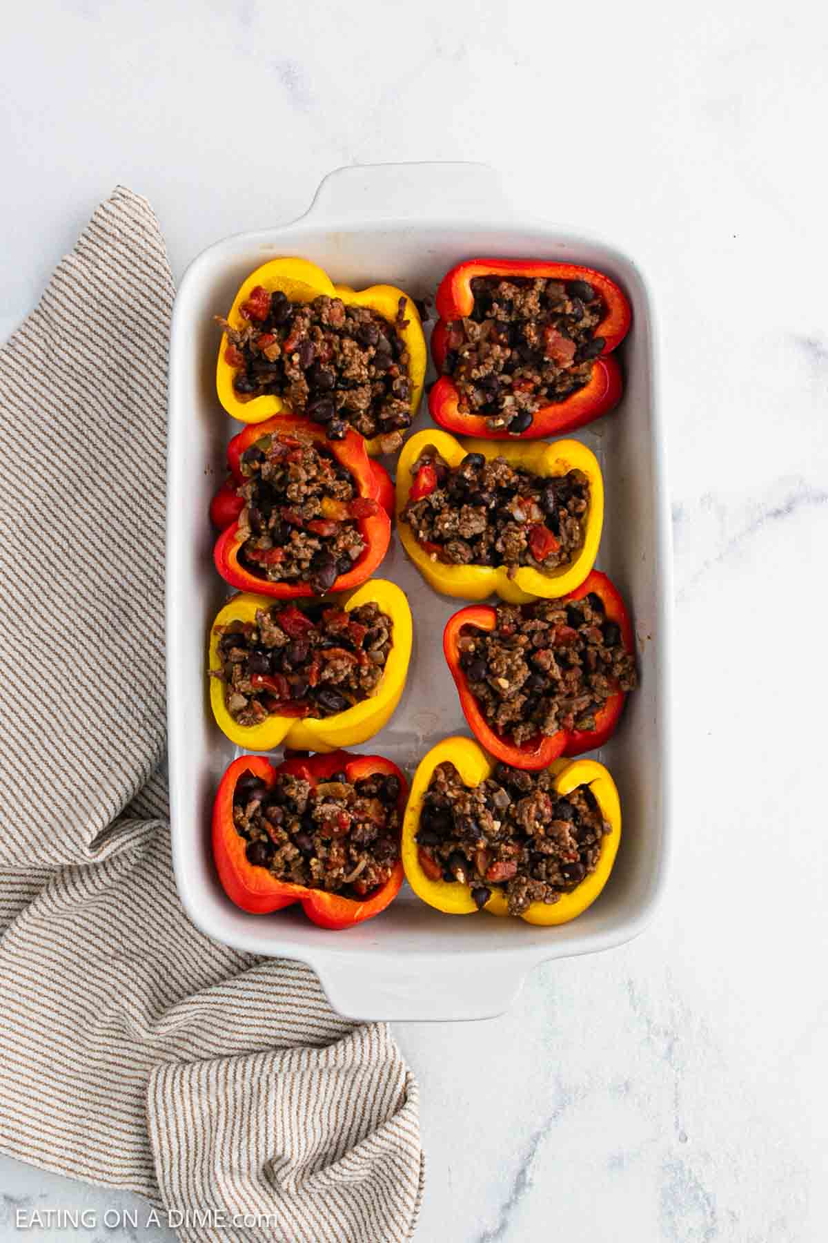 A white baking dish containing halved red and yellow bell peppers stuffed with a flavorful ground meat mixture, these Taco Stuffed Peppers rest on a white surface next to a striped kitchen towel.