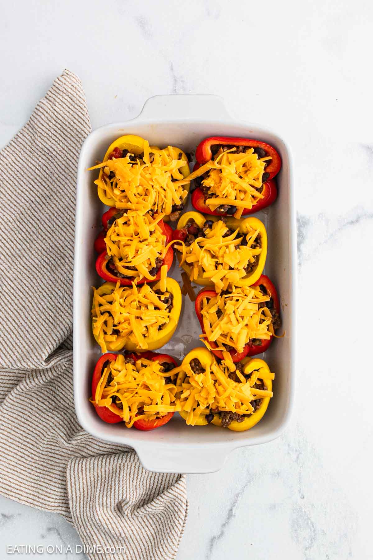 A white baking dish filled with taco stuffed peppers—halved red and yellow bell peppers packed with savory filling and topped with shredded cheddar cheese—sits on a white marble surface next to a striped cloth napkin.
