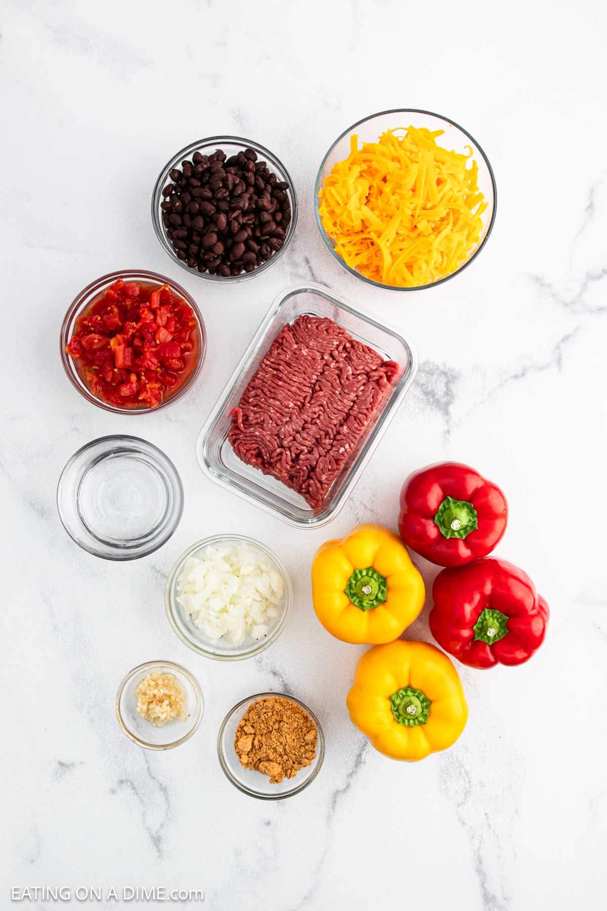 Flat lay of ingredients for Taco Stuffed Peppers on a white surface: ground beef, shredded cheddar cheese, black beans, diced tomatoes, red and yellow bell peppers, chopped onion, minced garlic, taco seasoning, and water. Perfect for a tasty taco recipe!.