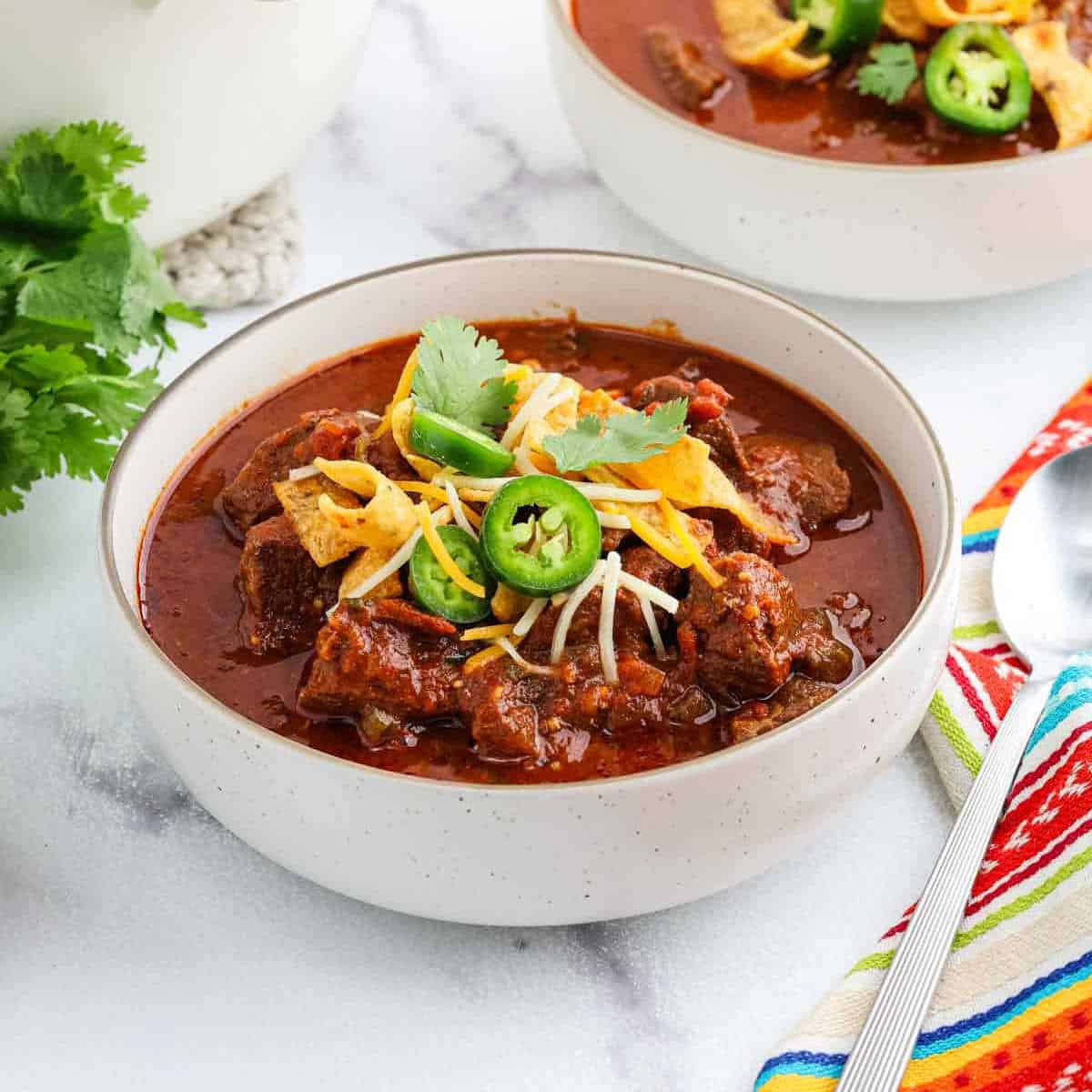A bowl of chili topped with shredded cheese, sliced jalapeรฑos, tortilla strips, and cilantro sits on a white surface with a spoon, colorful napkin, and another bowl in the background.
