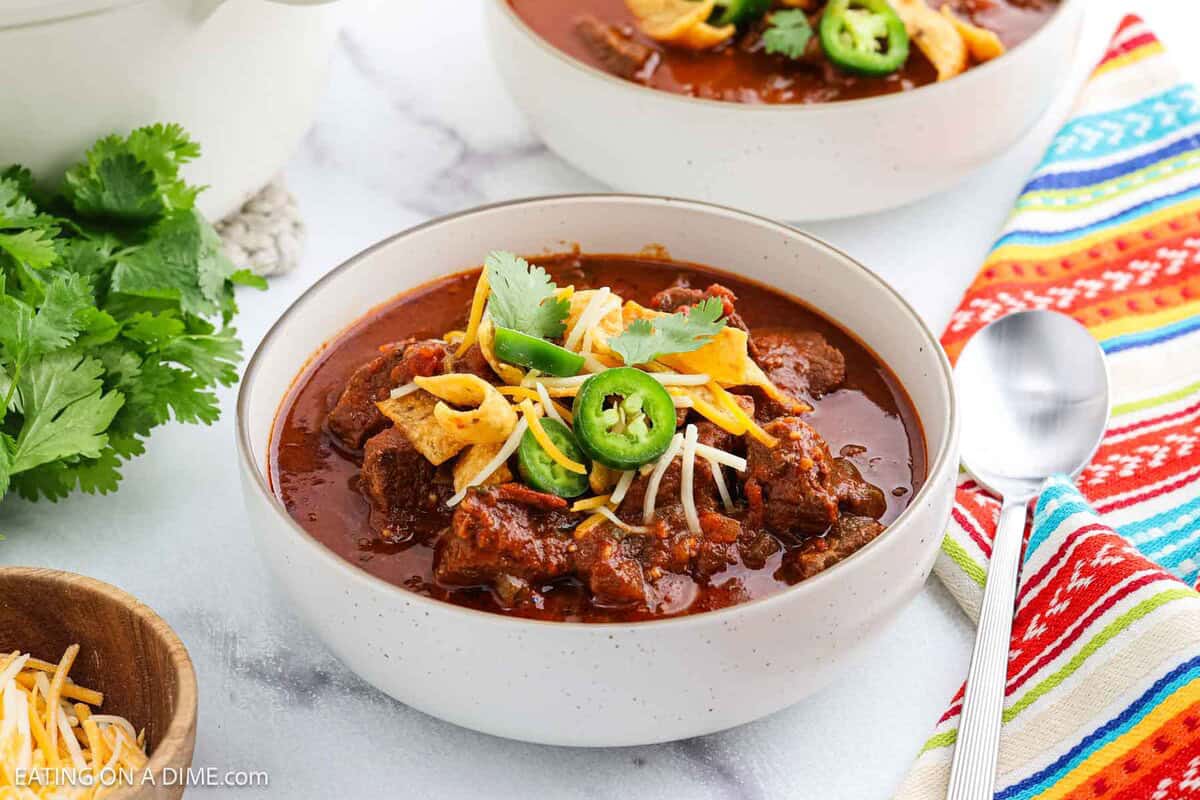 A bowl of chili topped with shredded cheese, cilantro, jalapeรฑo slices, and corn chips sits on a white surface next to a colorful striped napkin, a spoon, and fresh cilantro. Another bowl of chili is in the background.