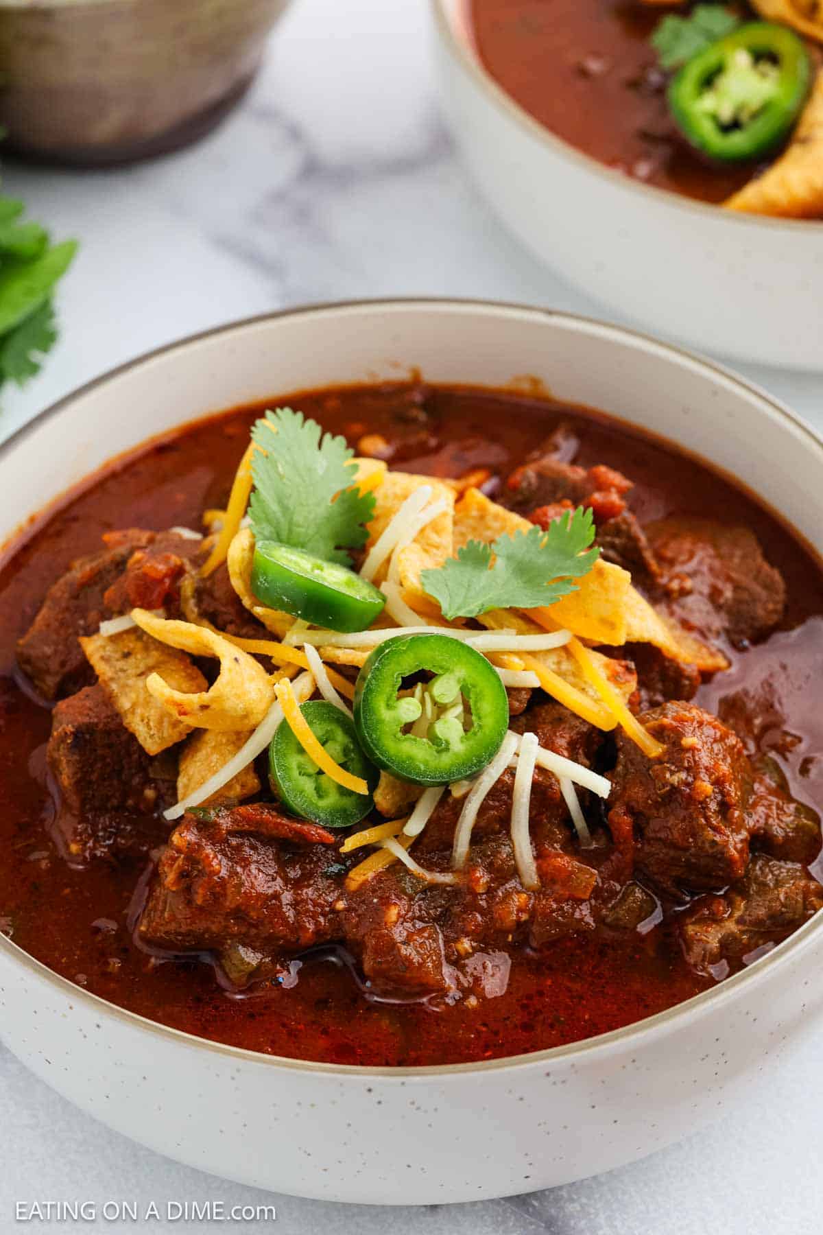 A bowl of chili topped with shredded cheese, Fritos corn chips, sliced jalapeรฑos, and fresh cilantro, served in a white bowl on a marble surface.
