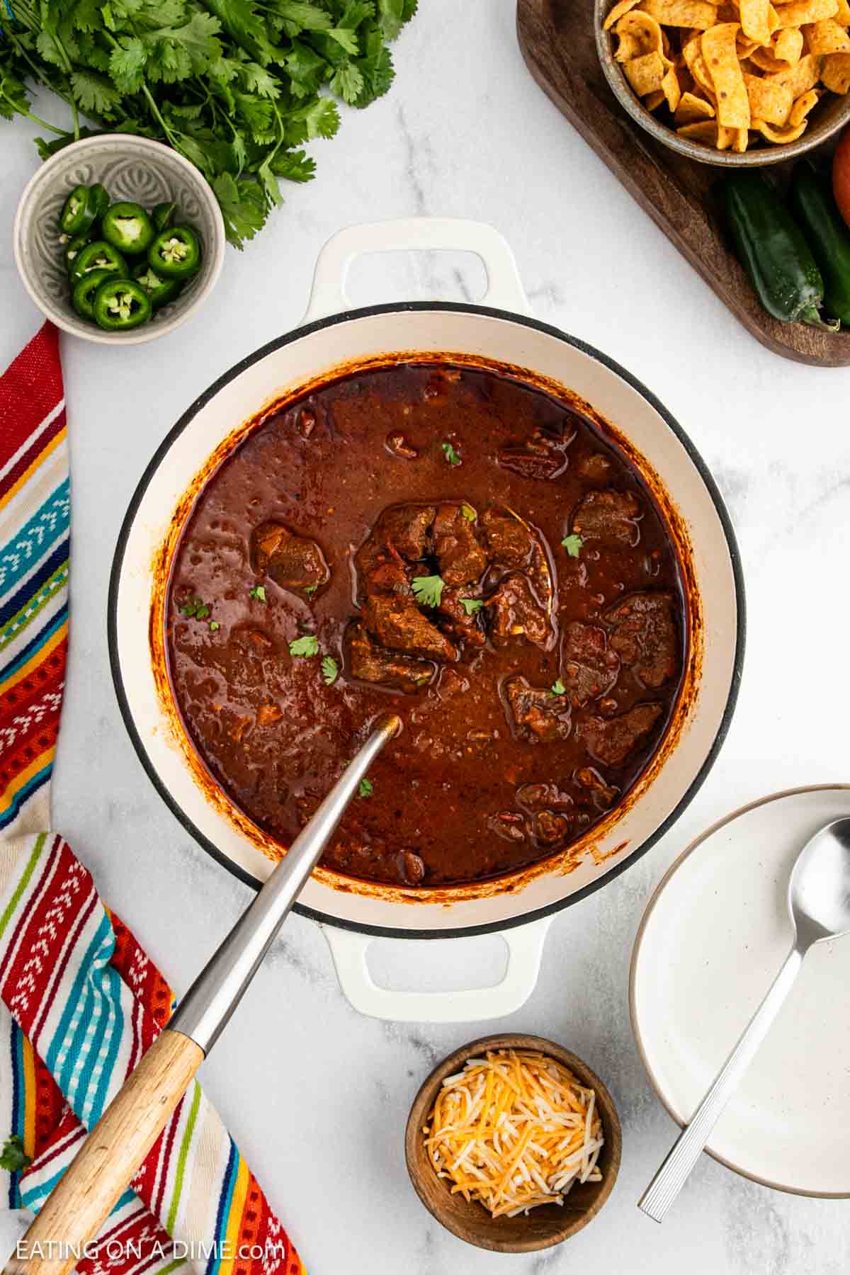 A pot of rich, red chili with chunks of beef, garnished with cilantro. Surrounding items include shredded cheese, sliced jalapeรฑos, corn chips, a spoon, and a colorful striped napkin on a white surface.