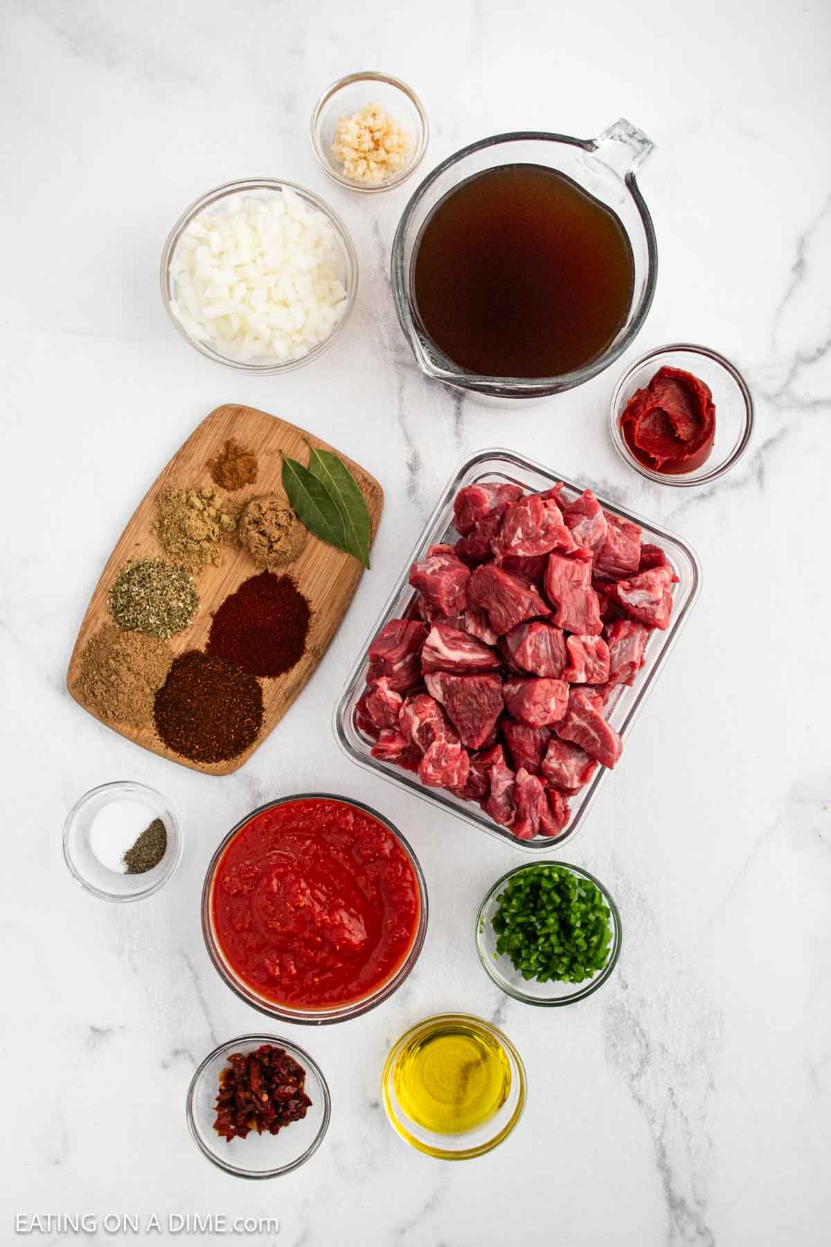 Top-down view of various ingredients for a stew, including cubed beef, diced onions, beef broth, tomato paste, crushed tomatoes, chopped peppers, minced garlic, spices, seasonings, and olive oil, arranged on a white surface.