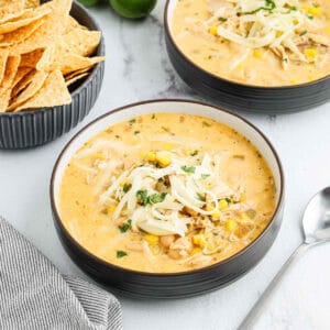 A bowl of creamy White Chicken Chili topped with shredded cheese and chopped herbs sits next to crispy tortilla chips, with a spoon and striped napkin on the side.