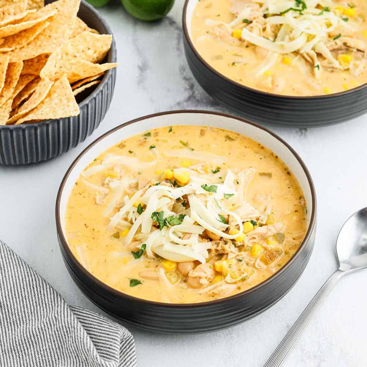 A bowl of creamy White Chicken Chili topped with shredded cheese and chopped herbs sits next to crispy tortilla chips, with a spoon and striped napkin on the side.