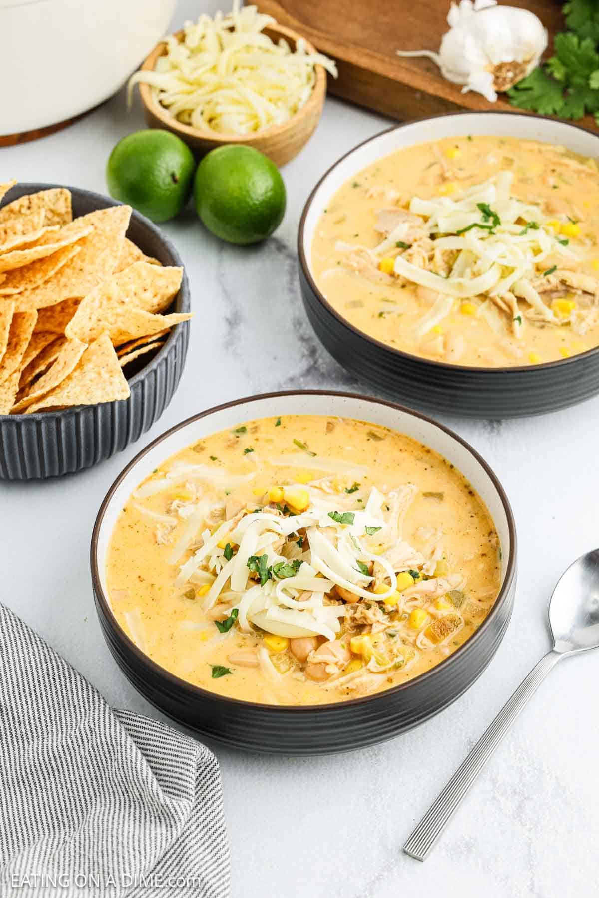 Two bowls of creamy White Chicken Chili topped with shredded cheese and cilantro, surrounded by tortilla chips, lime halves, extra cheese, garlic, and a spoon on a light-colored surface.